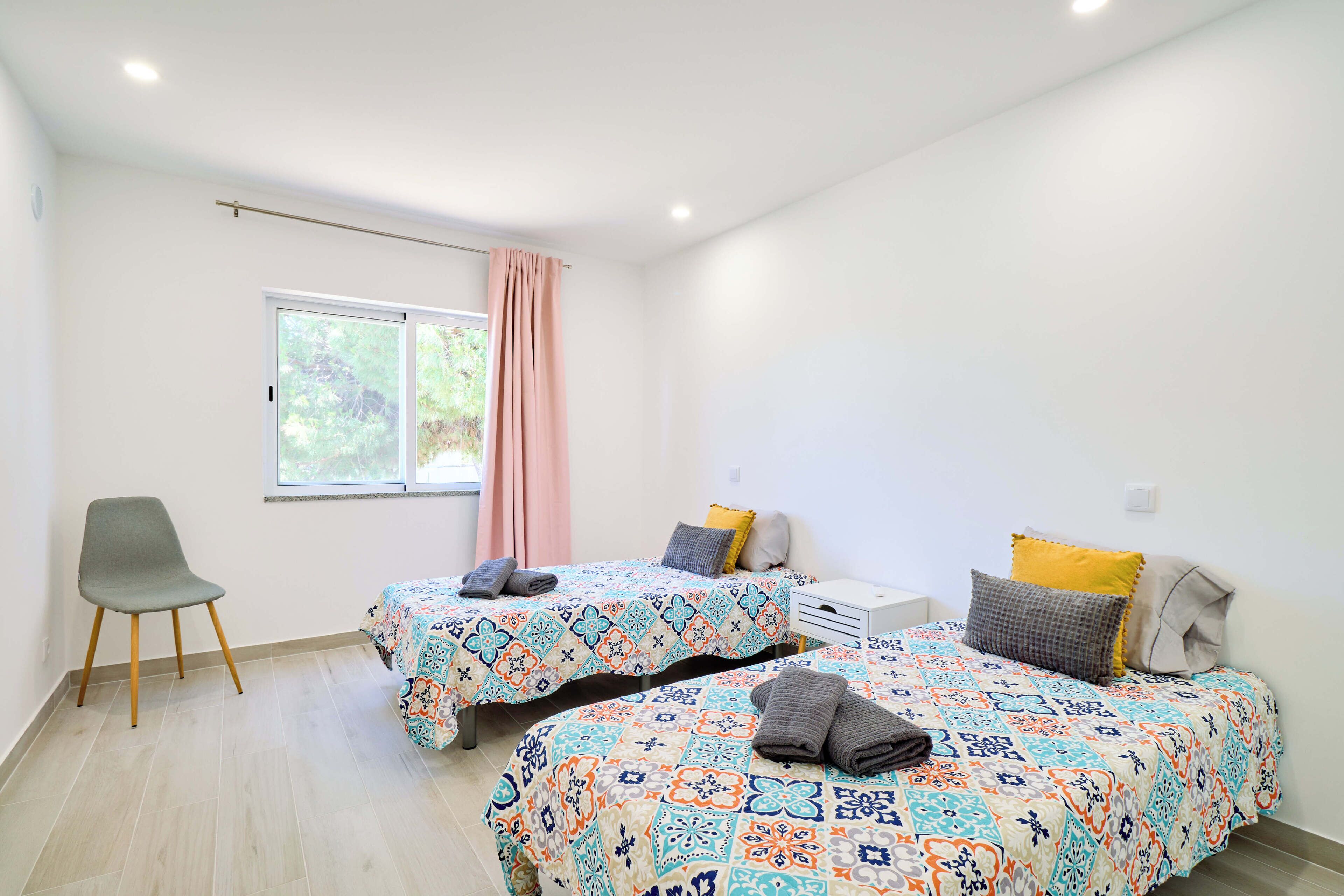 Una habitación con dos camas, una silla, un ventanal con cortinas y una mesita de noche blanca.
