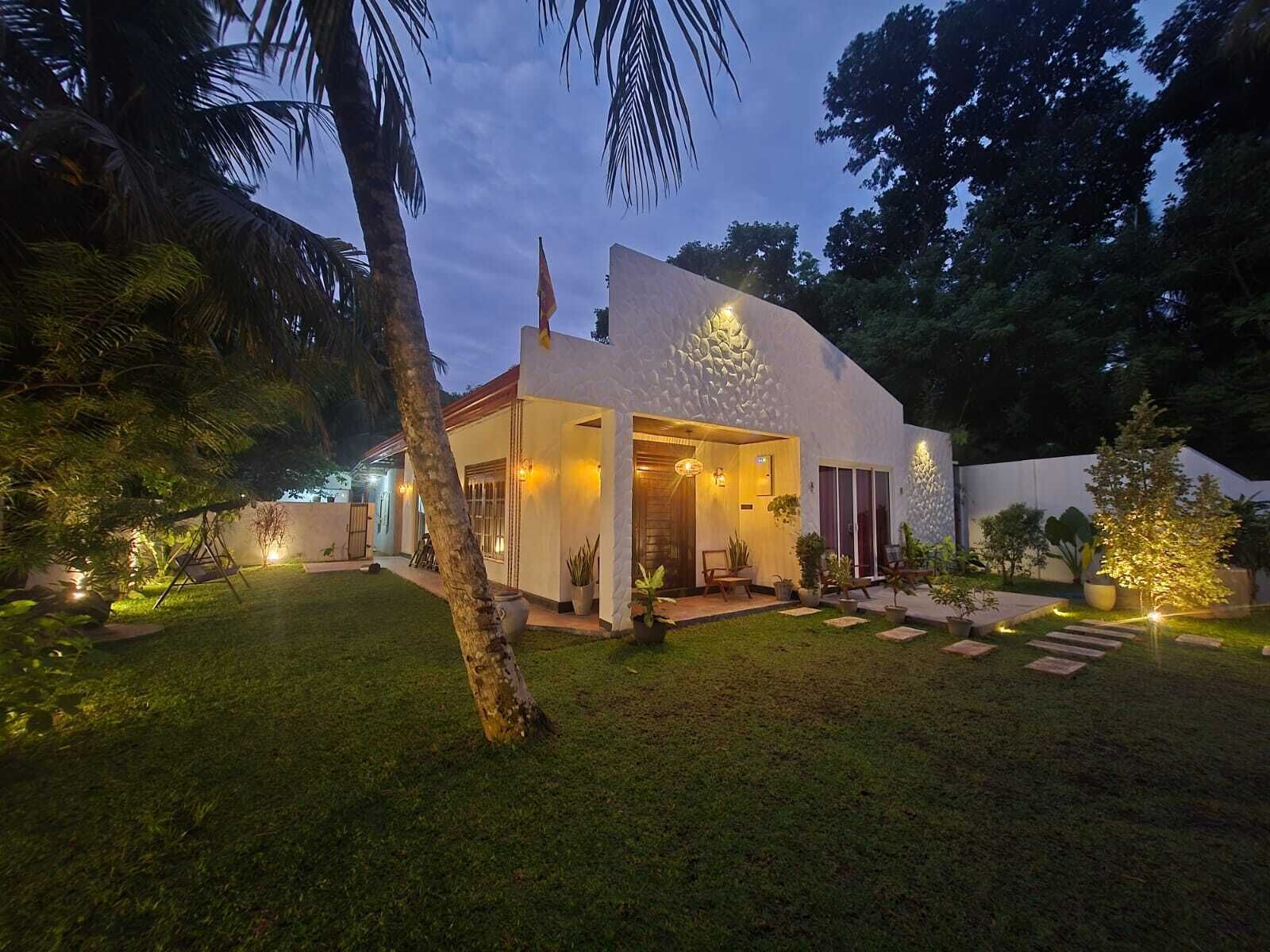 Une maison moderne avec une pelouse bien entretenue, un espace extérieur aménagé pour s’asseoir et un grand arbre au premier plan.