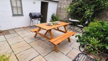 A patio with a wooden table and benches, a barbecue grill, and a small white building with a window.