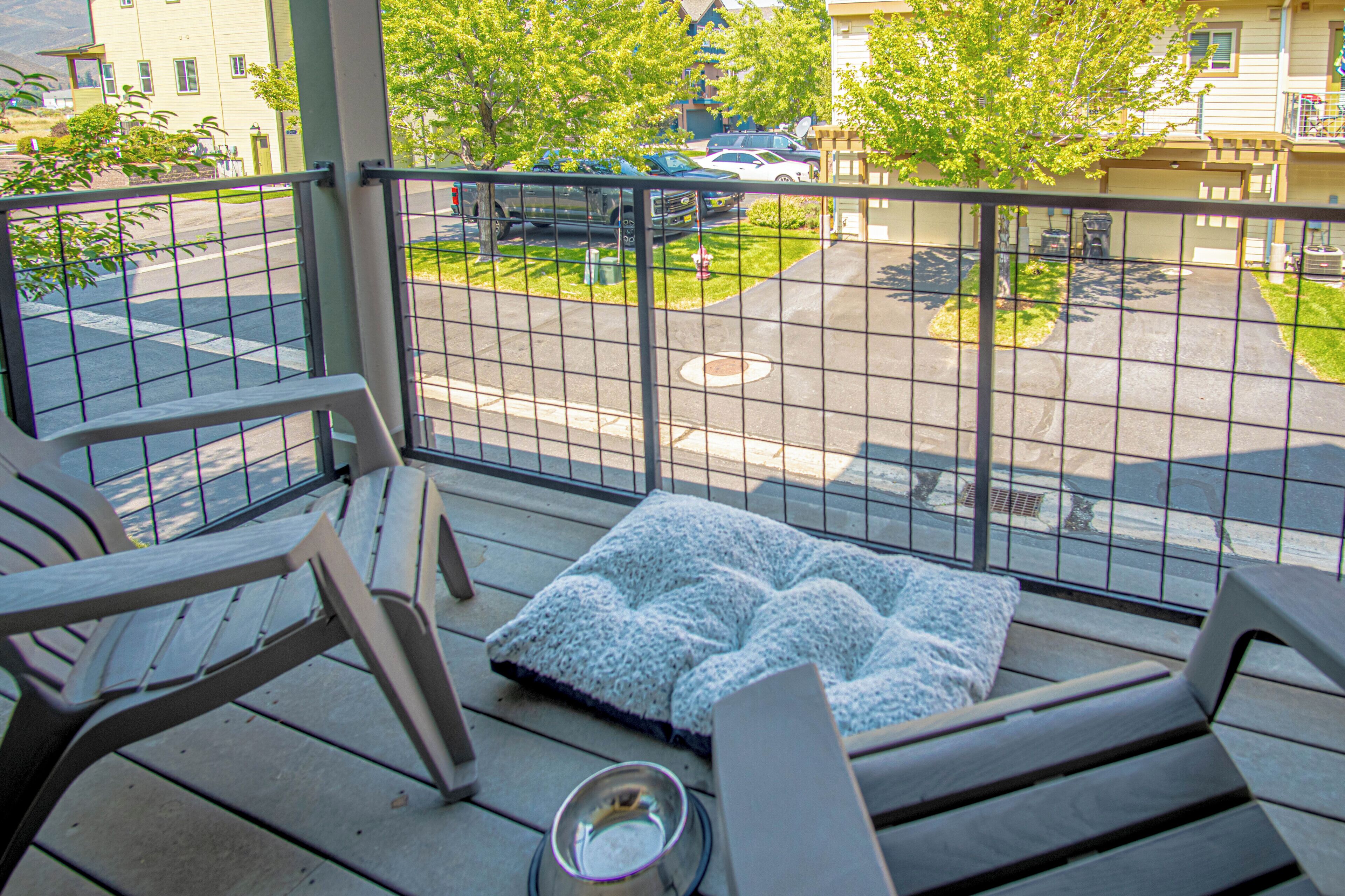 A balcony with a metal railing, a cushioned seating area, and a view of a street with parked cars and buildings.