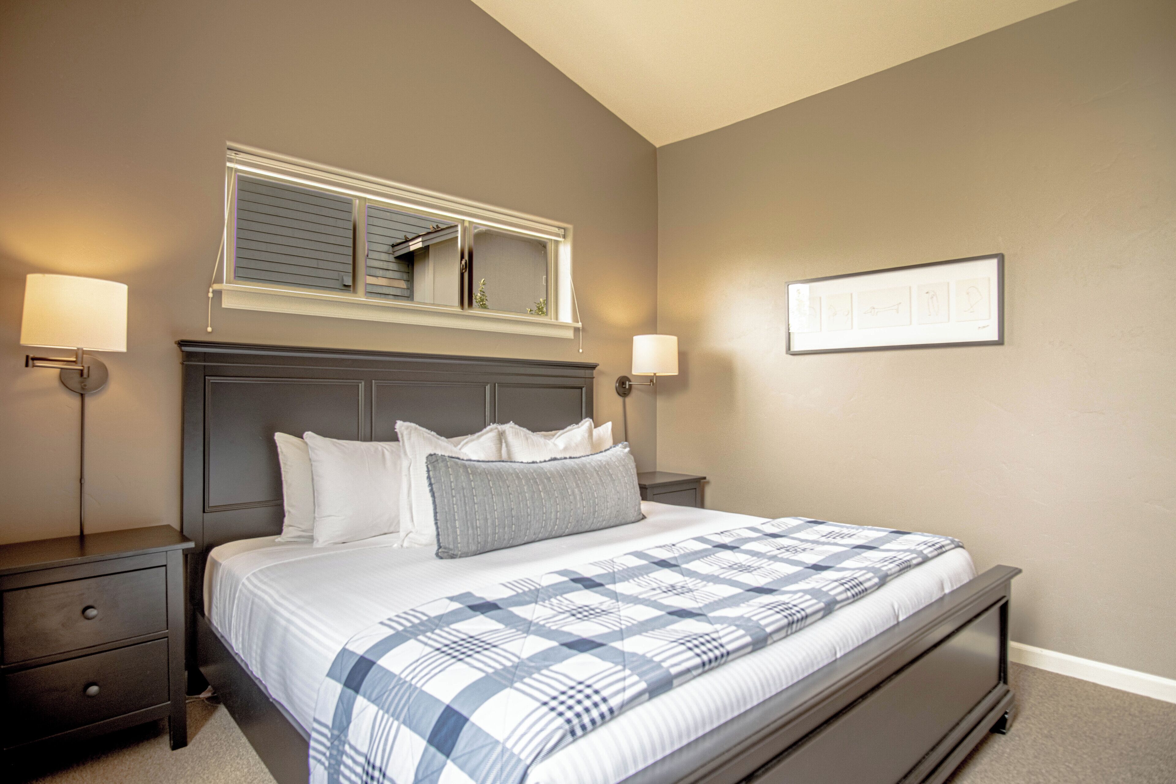 Un dormitorio con cama, mesitas de noche, ventana con persianas y lámparas fijadas a la pared.