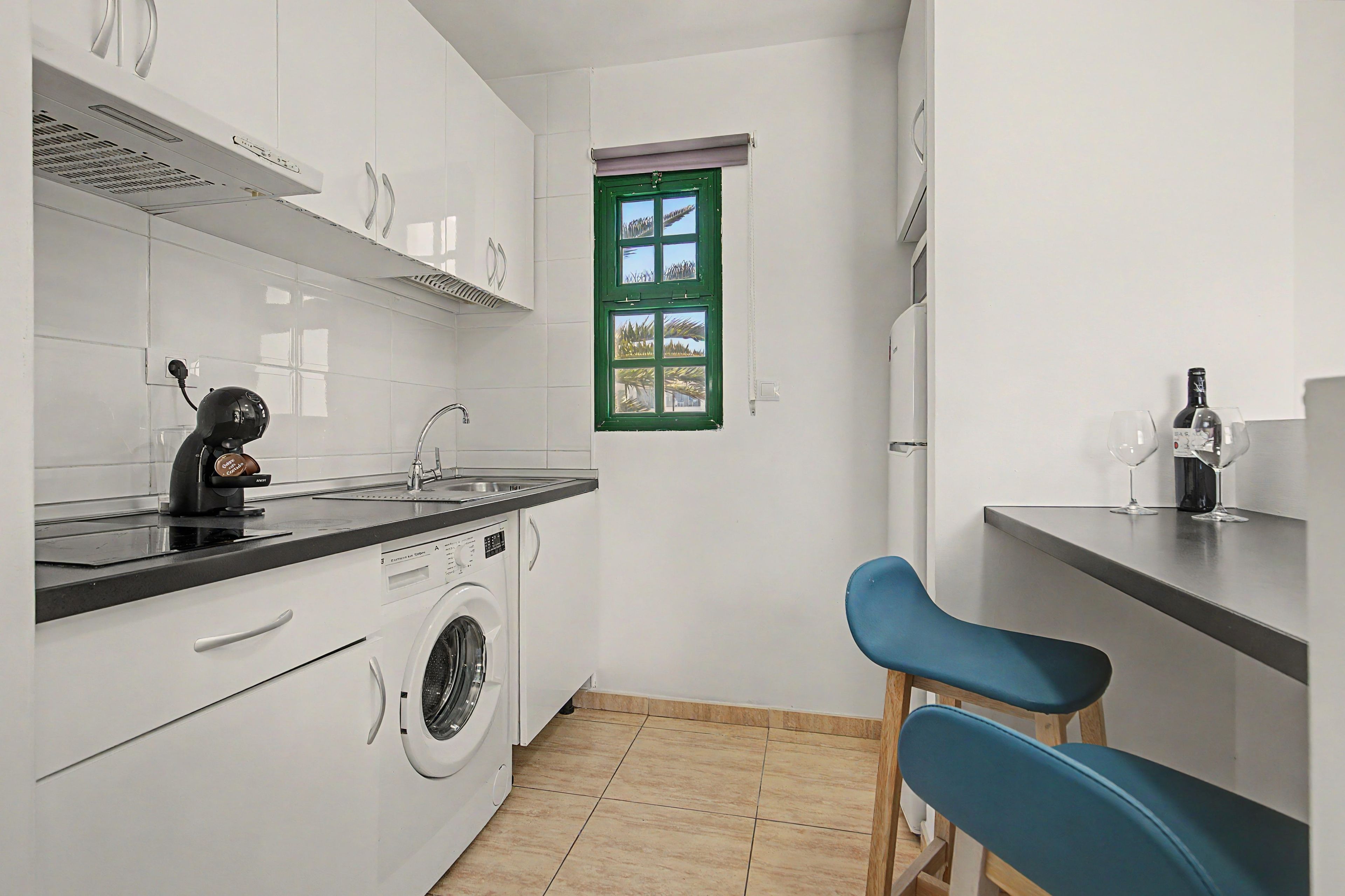A compact kitchen with a washing machine, a coffee machine, and a window with a green frame.
