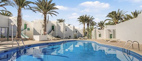 A swimming pool with clear blue water, surrounded by white buildings and palm trees.