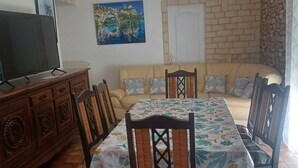 A dining area with a table set for four, surrounded by chairs with a floral tablecloth. A television is on a wooden cabinet to the left. - Les Oliviers Vacation Rental (pleubian)