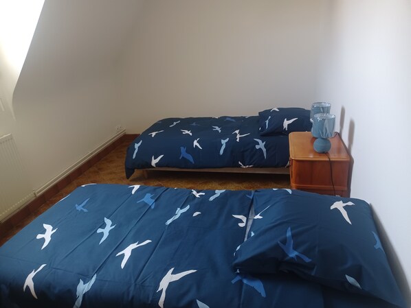 A bedroom with two beds, each with a blue bedspread featuring white bird patterns, a wooden nightstand with a blue lamp, and a radiator on the wall. - Les Oliviers Vacation Rental (pleubian)