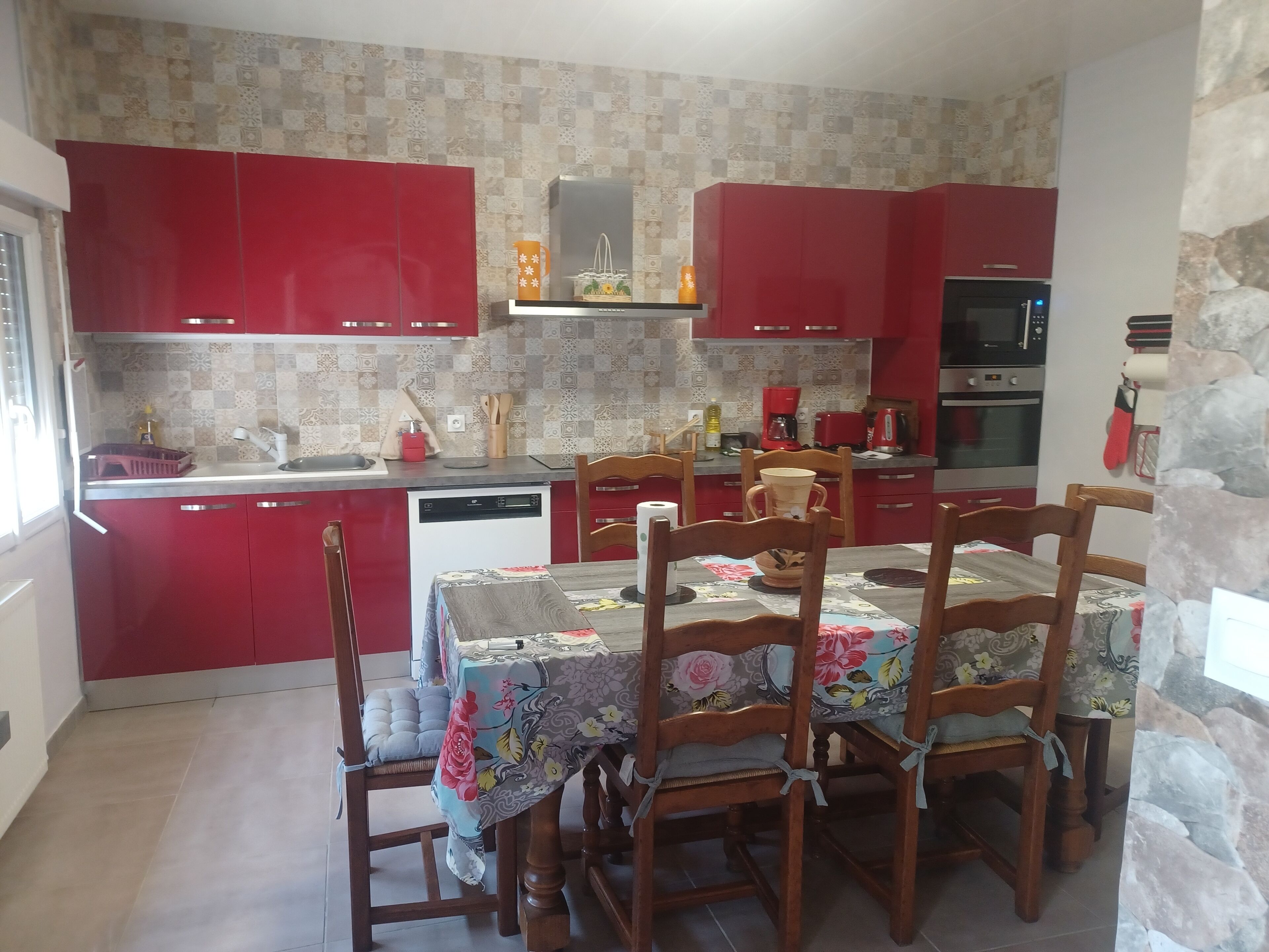 A kitchen with red cabinets, a dining table with floral tablecloth, and a stone wall.
