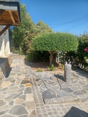 A stone-paved patio with a stone pillar, a wooden structure, and a well-maintained garden with various plants and a tree. - Les Oliviers Vacation Rental (pleubian)