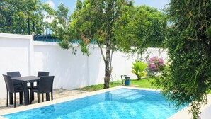 Una piscina rectangular con un diseño de mosaicos, rodeada por un muro blanco y un patio empedrado con una mesa y sillas.