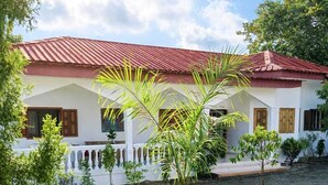 Una casa con techo rojo, paredes blancas y un pequeño jardín con una palmera.