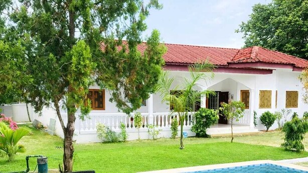 Una piscina con zona de borde blanca, una mesa con sillas y una casa de techo rojo al fondo.