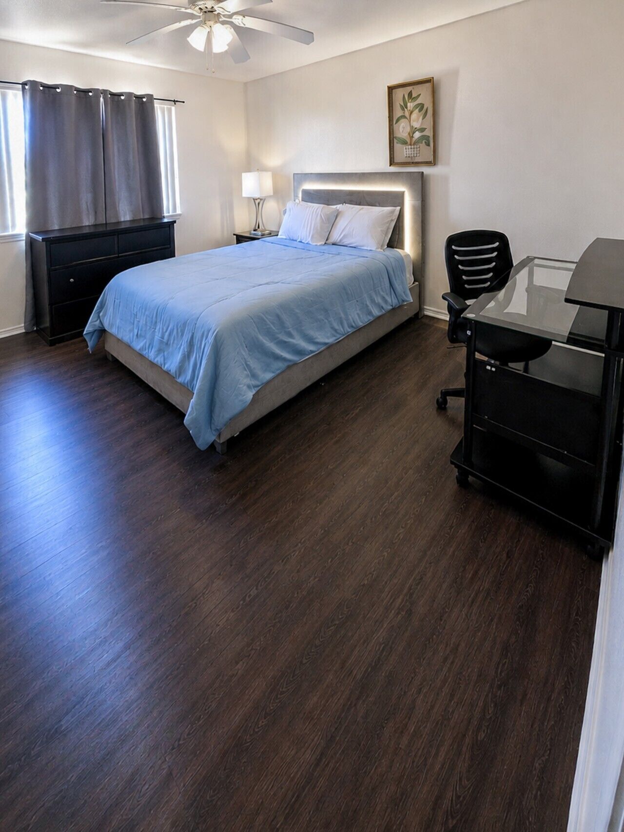 A bedroom with a bed, a desk, a chair, a ceiling fan, and a window with curtains.