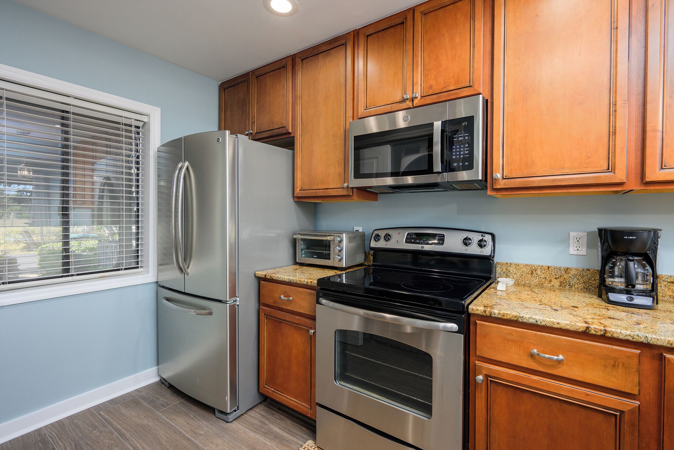 A kitchen with stainless steel appliances, wooden cabinets, a window with blinds, and a granite countertop.