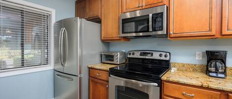 A kitchen with stainless steel appliances, wooden cabinets, a window with blinds, and a granite countertop.