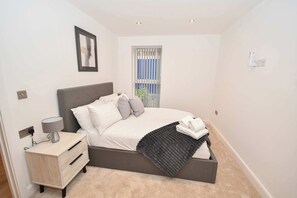 A bedroom with a bed, bedside table, lamp, and a window with blinds. - Your Home Away From Home in Central Leeds LS9 (Leeds)