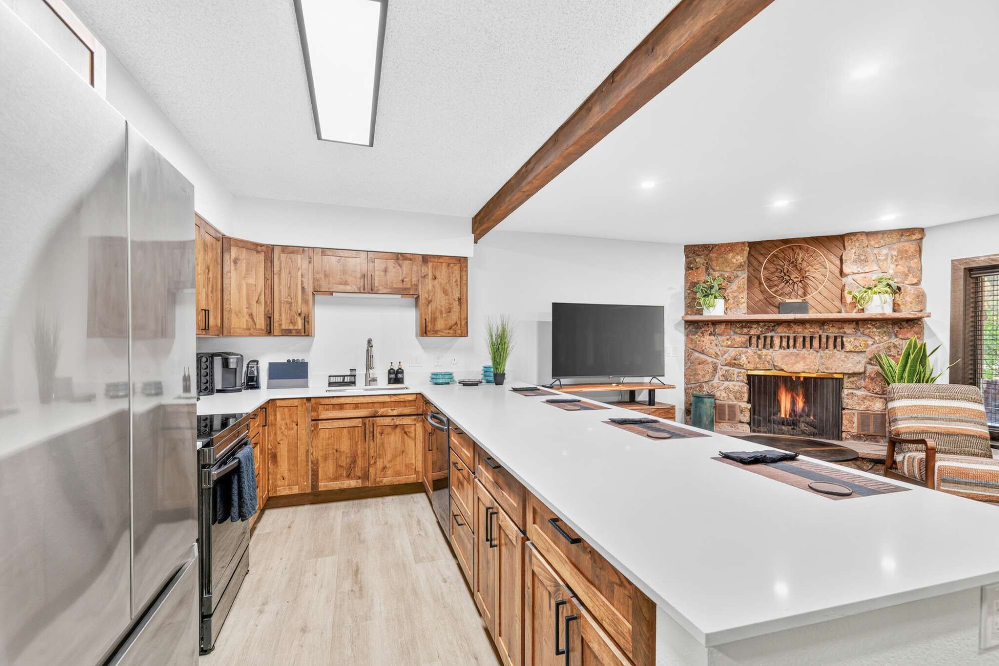 A modern kitchen with a large island, wooden cabinets, a stone fireplace, and a flat-screen TV.