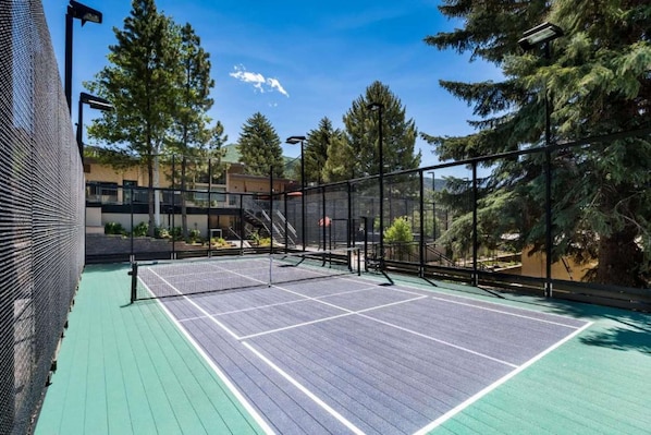 Een buitenterrein voor tennis met een net, omringd door een metalen hek en bomen.
