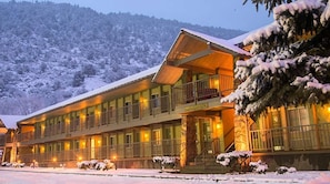 Ett hotell med en balkong och flera fönster, omgivet av snötäckta träd och en bergig bakgrund.