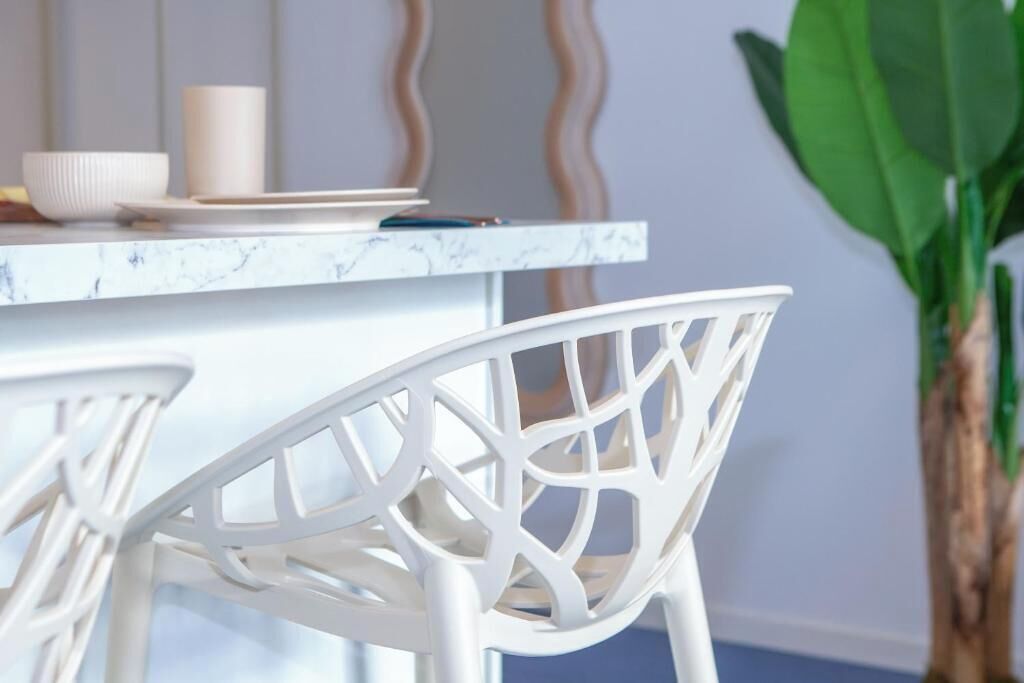 A white chair with a geometric design, a white table with a marble-like surface, and a potted plant in the background.