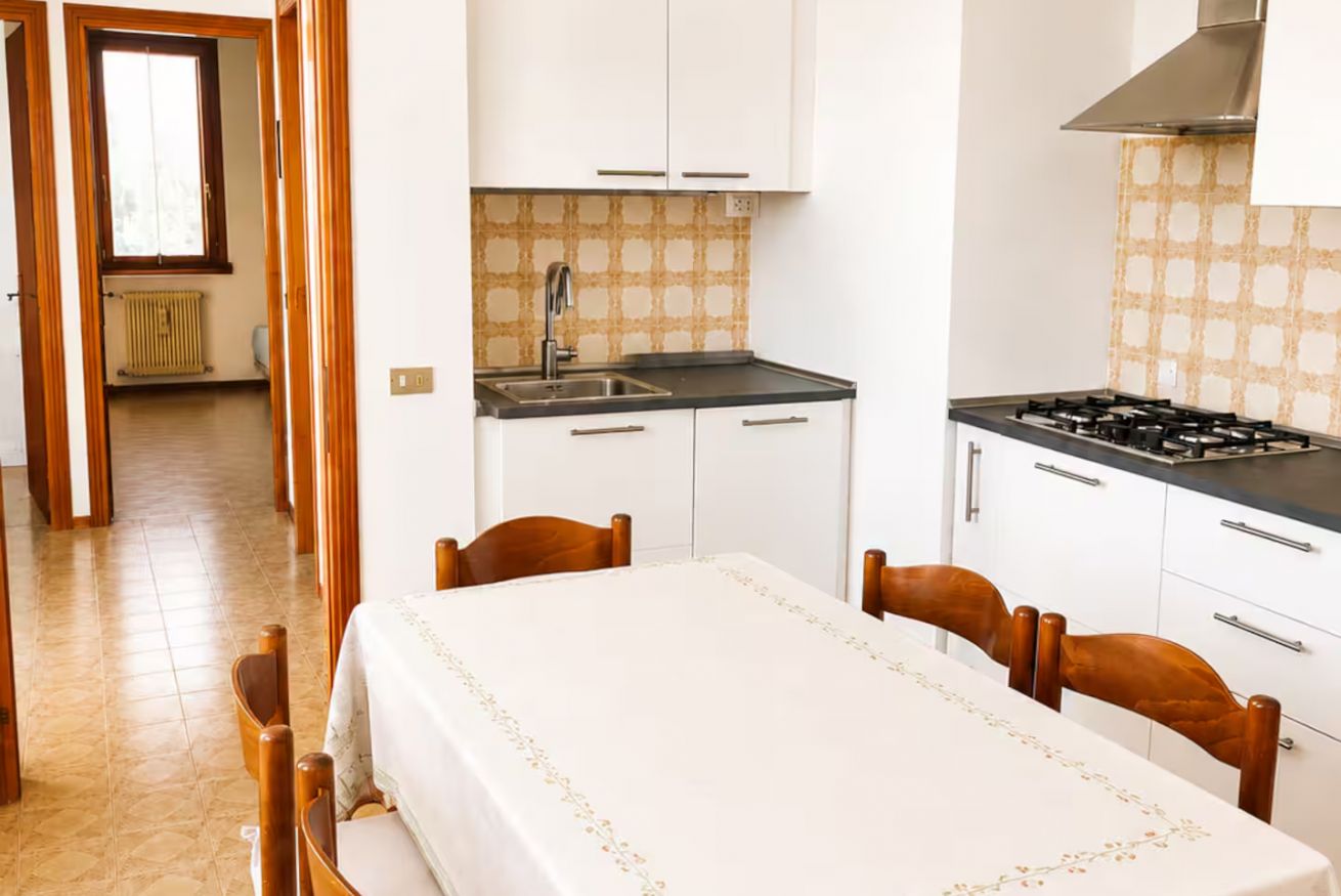 A compact kitchen with a stove, sink, and white cabinets. A dining table with chairs is positioned in front of the kitchen.