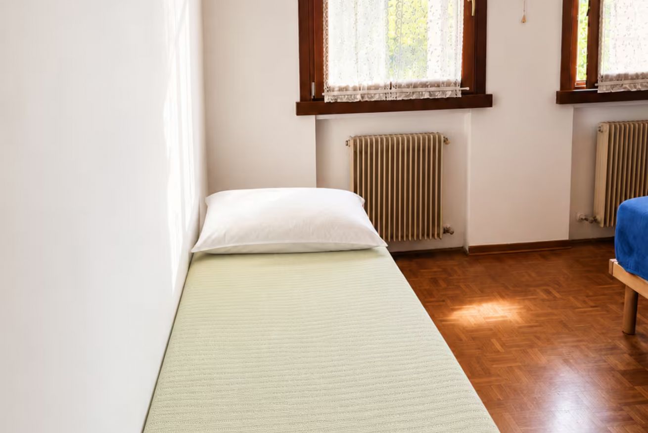 A single bed with a white pillow and a light green mattress cover in a room with wooden flooring and two radiators.