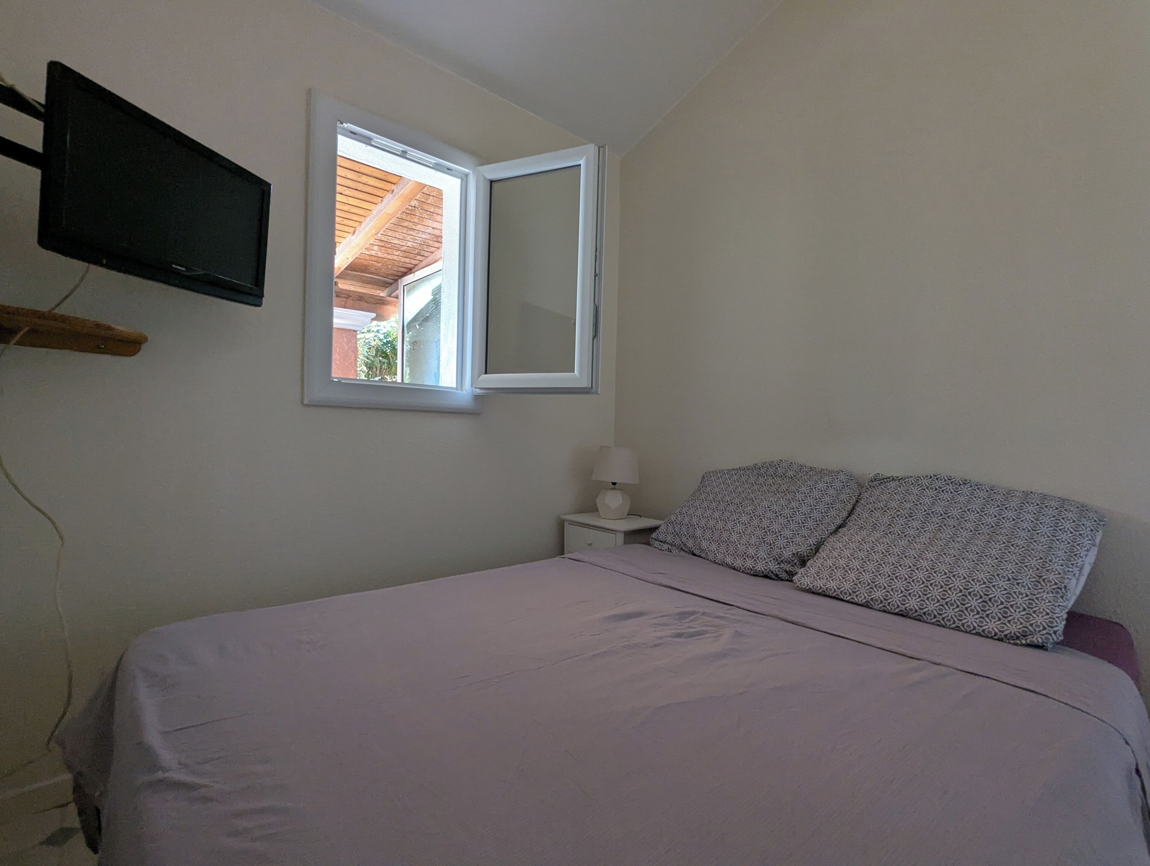Ein Schlafzimmer mit Bett, Nachttisch mit Lampe, Wand-Fernseher und Fenster.
