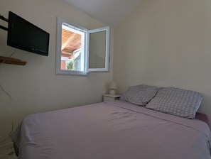 A bedroom with a bed, a bedside table with a lamp, a TV mounted on the wall, and a window. - Detached house with outdoor garden for rent in a gated community with a pool (Loupian)