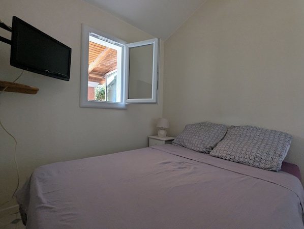 A bedroom with a bed, a bedside table with a lamp, a TV mounted on the wall, and a window. - Detached house with outdoor garden for rent in a gated community with a pool (Loupian)