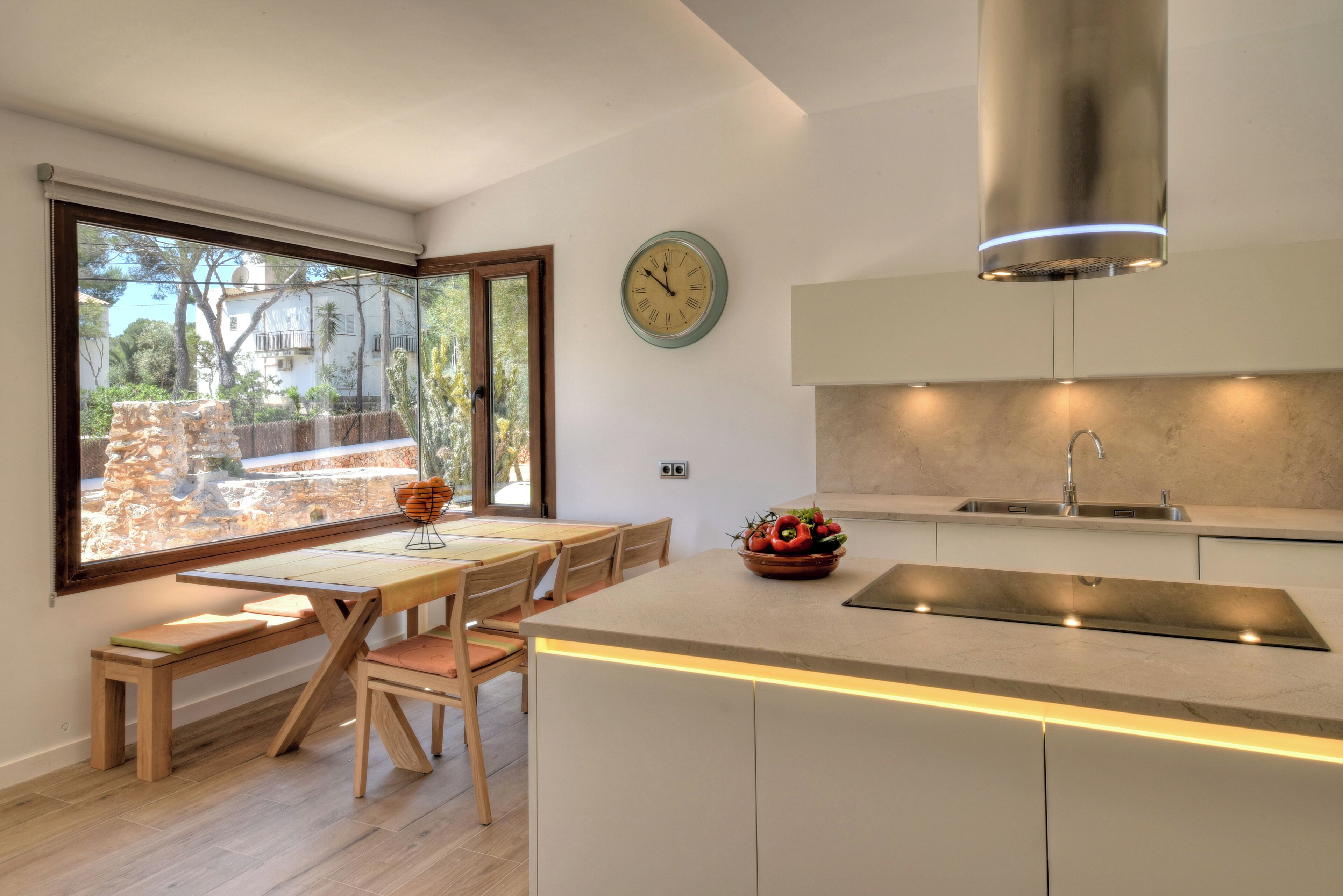 A modern kitchen with a wooden dining table, a large window, and a clock on the wall.