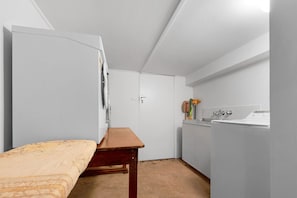 A compact room with a bed, a small table, a washing machine, and a door leading to another room.