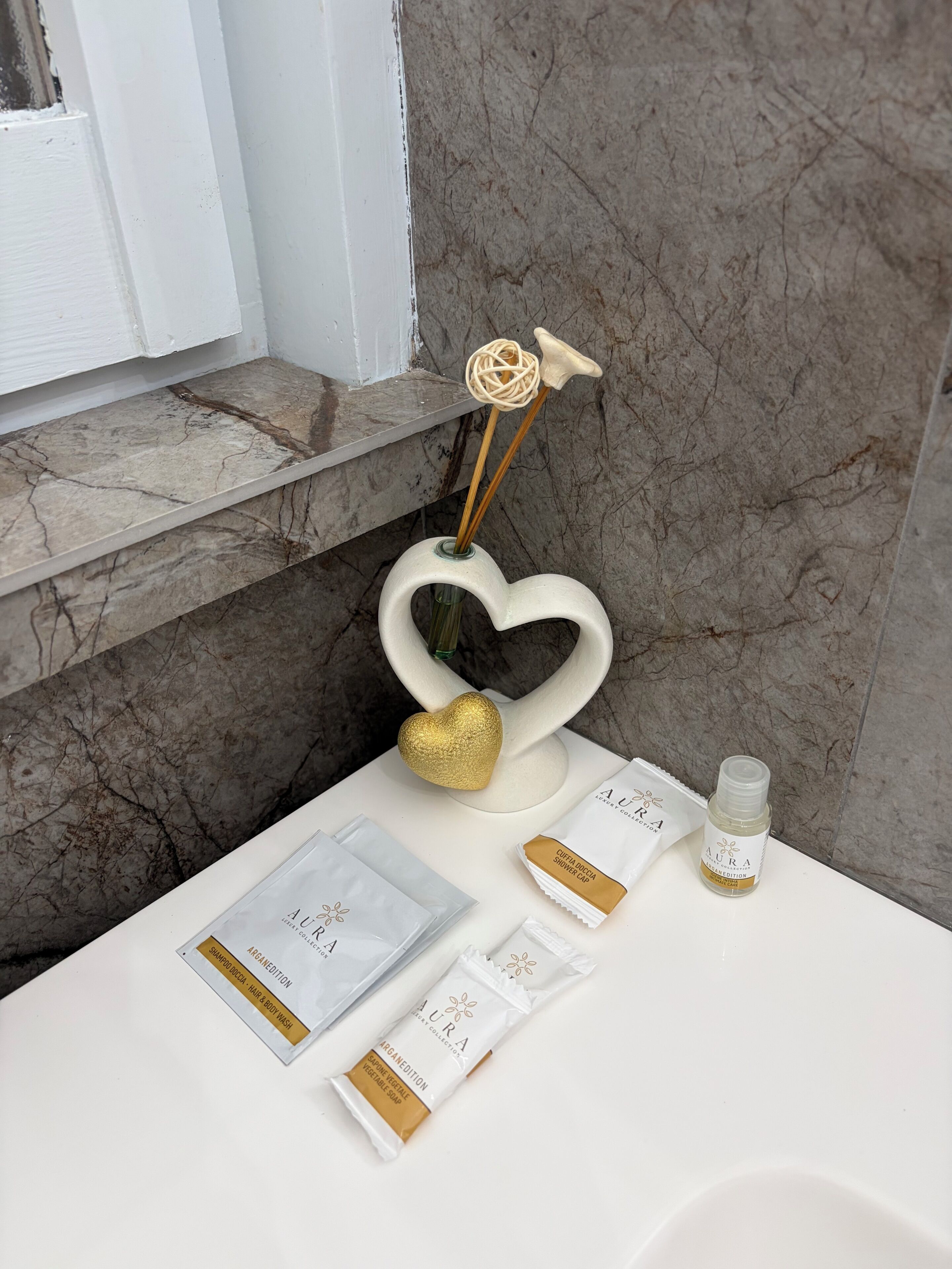 Un meuble-lavabo de salle de bain avec un plan de toilette en marbre, un porte-savon blanc en forme de cœur et divers articles de toilette, notamment un bonnet de douche et un flacon de lotion.
