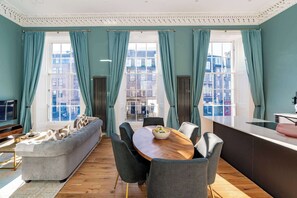 Ein modernes Wohnzimmer mit Sofa, Esstisch und Stühlen, großen Fenstern mit Vorhängen und Blick auf Gebäude.
