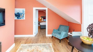Una habitación con piso de madera, un sillón azul, una mesa pequeña con un frutero y un cuadro en la pared.