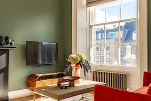 Ein Wohnzimmer mit an der Wand befestigtem Fernseher, einer Holzkommode, einem Tisch mit Blumenvase und einem roten Sessel.