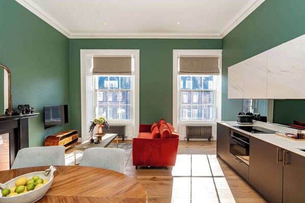 A modern living room with a red sofa, a wooden dining table with a bowl of fruit, and a kitchen area with a large white cabinet. - Stevenson Suite - 1 Bed Luxury Apartment (Edinburgh)