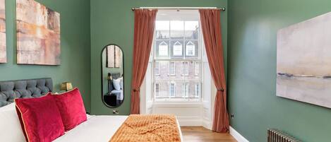 Un dormitorio con cama, un espejo, un radiador y vistas a edificios a través de una ventana.