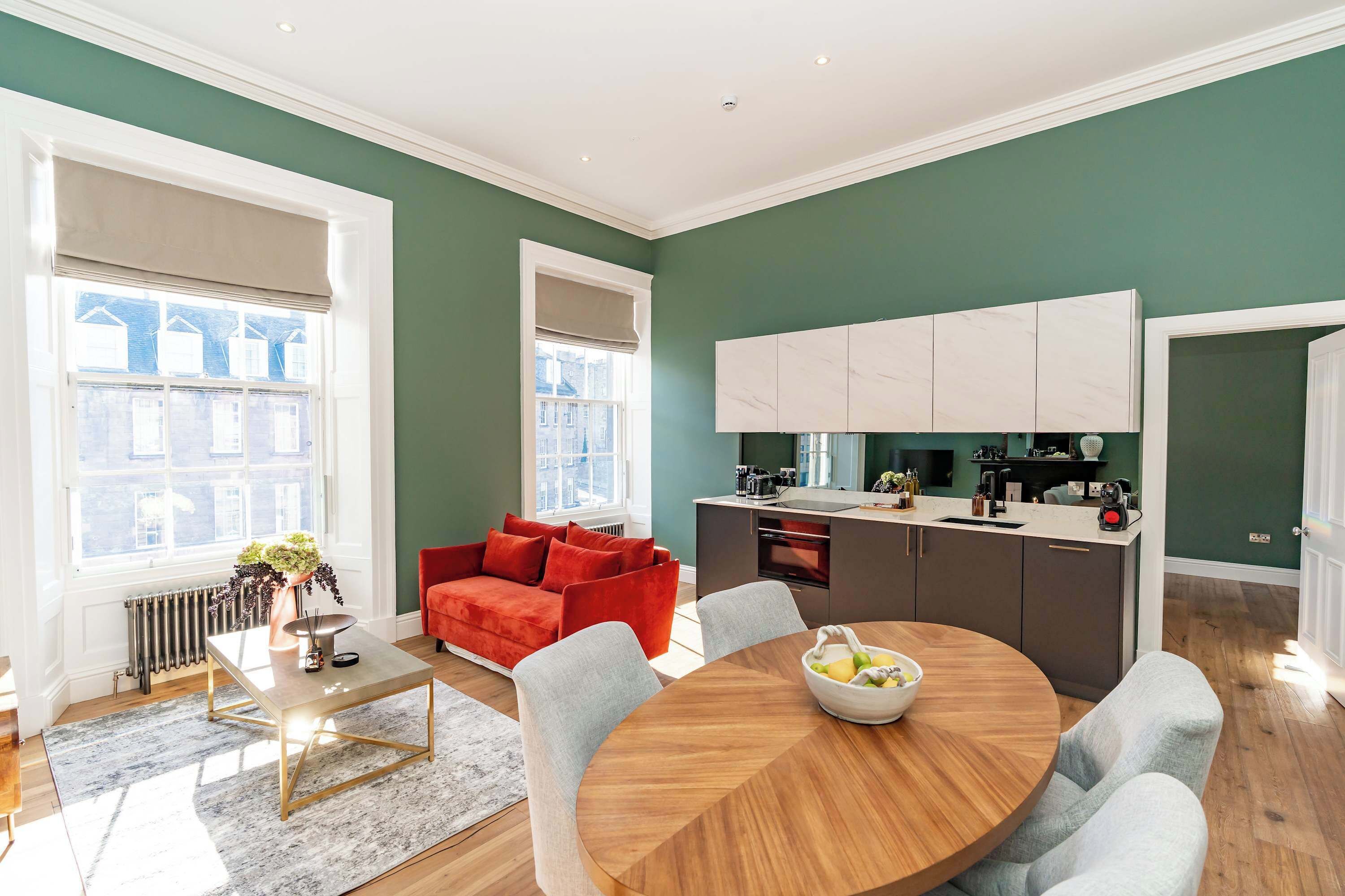 A modern living room with a red sofa, a round dining table, and a kitchen with white cabinets.