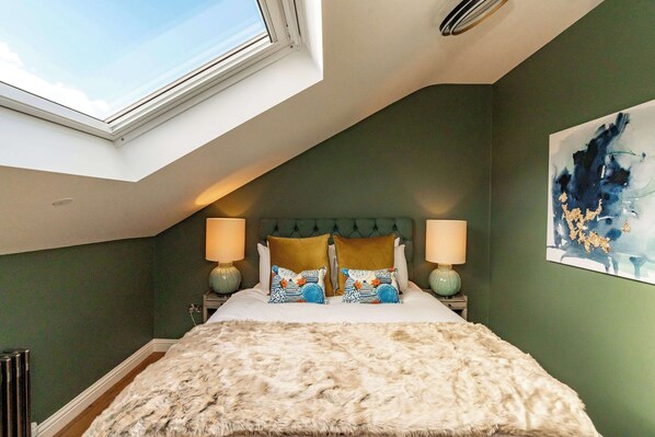Une chambre à coucher dotée d’une lucarne, d’une tête de lit capitonnée, de deux lampes de chevet, d’oreillers à motifs et d’un plaid en fourrure.