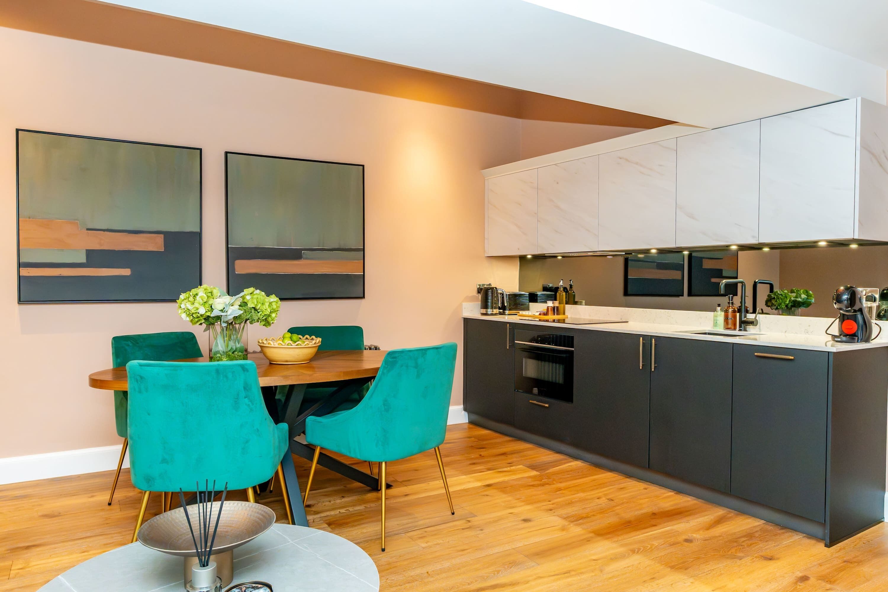 Une cuisine moderne avec des meubles de couleur sombre, un plan de travail blanc et un coin repas agrémenté de chaises turquoise et d’une table en bois.