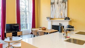 A modern living room with a TV, dining table, and chairs. There is a fireplace, a kitchen area with a sink, and a large abstract painting on the wall. - 2 Bed Luxury Apartment (Edinburgh)