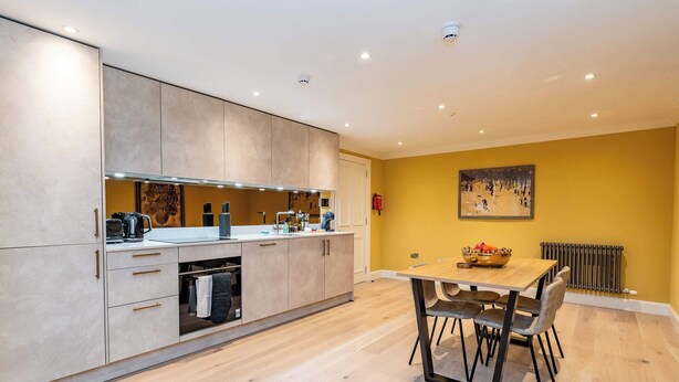 A modern kitchen with a dining area, featuring a wooden table and chairs, a built-in oven, and a painting on the wall. - Dragon Suites 1 Bed Luxury Apartment (Edinburgh)