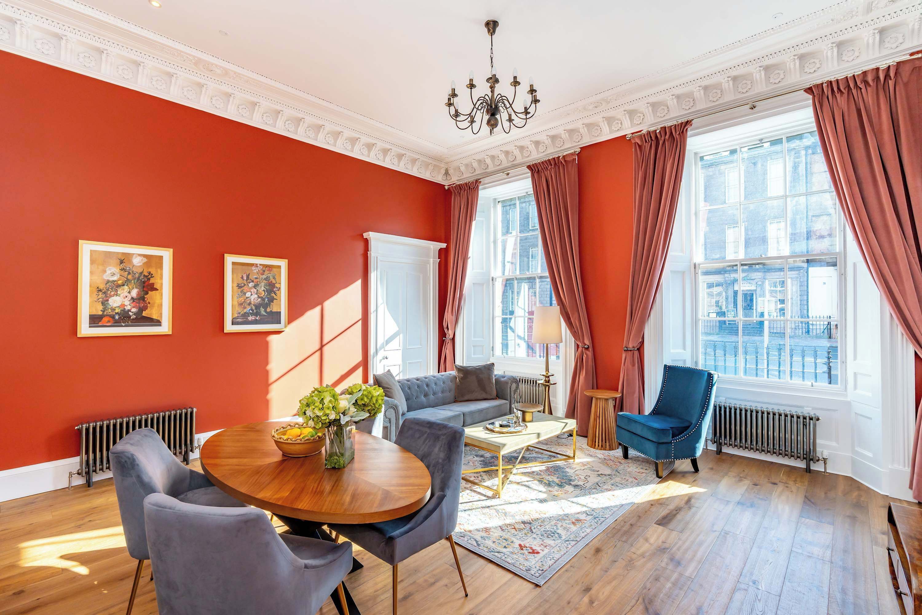 Una habitación con pared roja, una lámpara de araña, un sofá, un sillón azul, una mesa redonda de madera y dos cuadros con motivos florales.