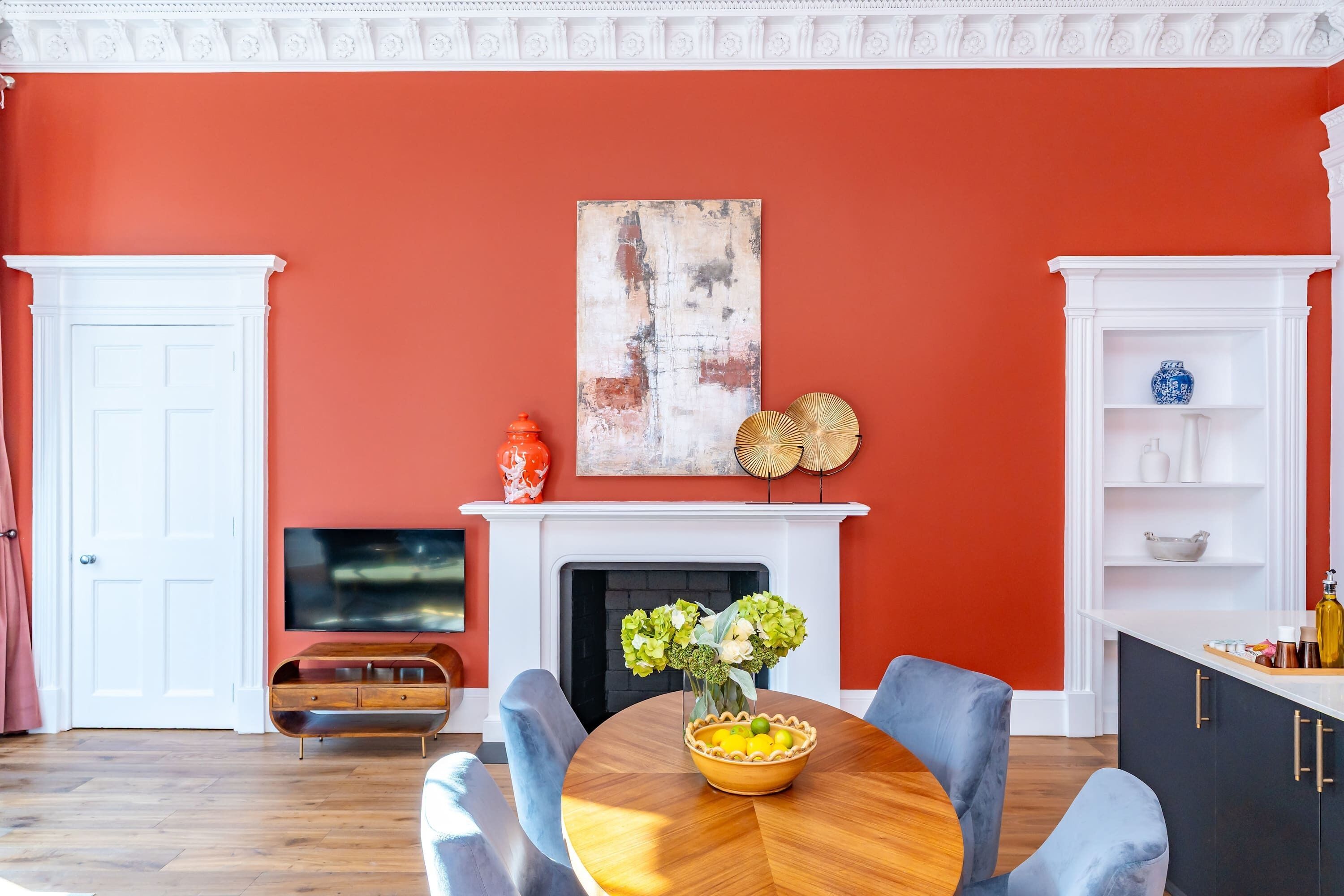 A living room with a fireplace, a television, a dining table with chairs, and a red accent wall.