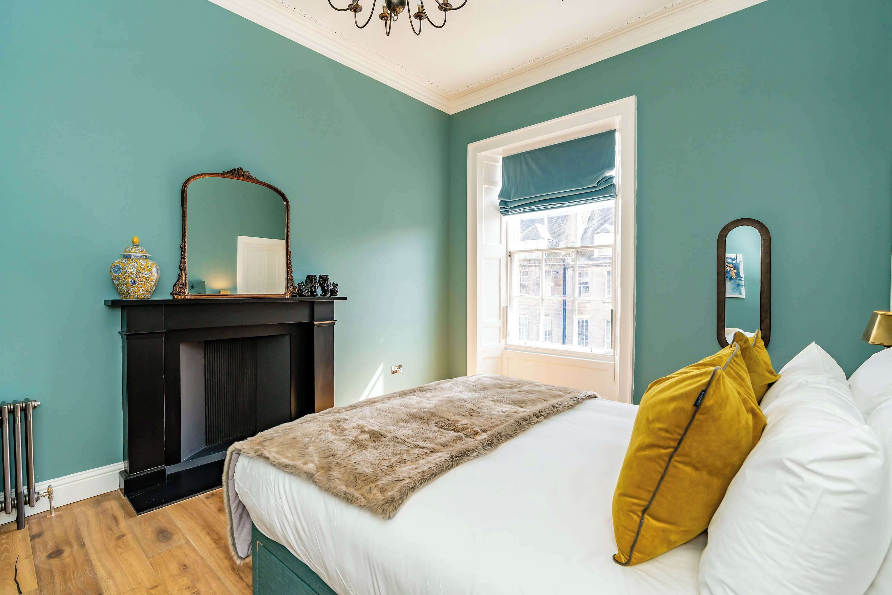 Un dormitorio con paredes color turquesa, una chimenea, una cama con almohadas blancas y mostaza, y una ventana con vista a edificios.