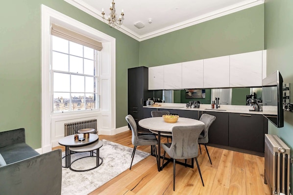 A modern kitchen with a round dining table, grey chairs, and a view through a large window. - Mackintosh Suite - 1 Bed Luxury Apartment (Edinburgh)