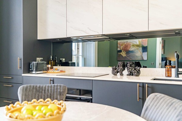 A modern kitchen with grey cabinets, a marble countertop, and a dining area with a round table. - Mackintosh Suite - 1 Bed Luxury Apartment (Edinburgh)