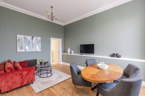 A living room with a red sofa, a round wooden dining table with chairs, a flat-screen TV mounted on the wall, and a chandelier. - Latta Suite - 1 Bed Luxury Apartment (Edinburgh)