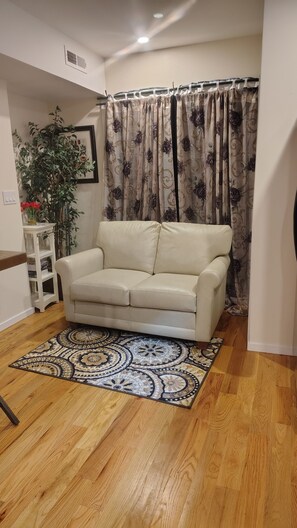 A living room with a beige sofa, a patterned rug, wooden flooring, a potted plant, and a curtain with a floral design. - Peaceful 2-bedroom house with AC in charming West Haven, near Yale & VA hospital (West Haven)