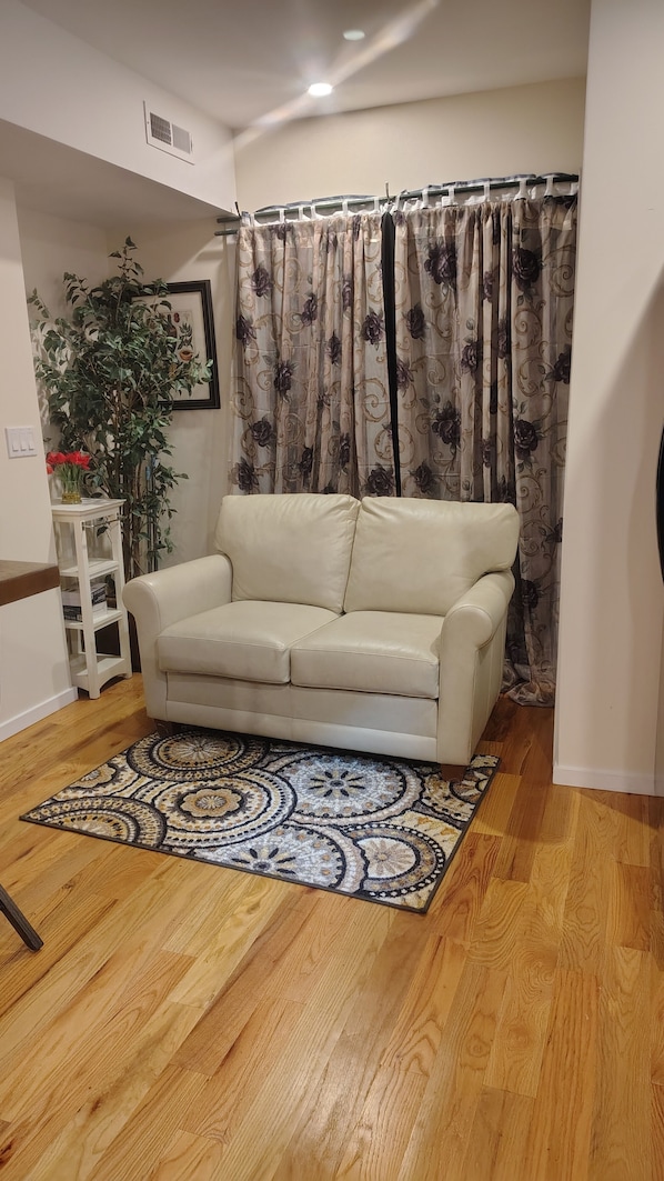 A living room with a beige sofa, a patterned rug, wooden flooring, a potted plant, and a curtain with a floral design.