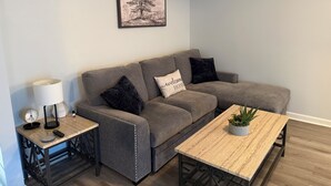 A living room with a grey sofa, two black cushions, a white lamp, a small table with a plant, and a framed picture on the wall. - Perfect Location for Gameday (Oxford)