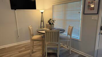 A dining area with a round table, two chairs, a wall-mounted TV, a floor lamp, and a window with blinds.